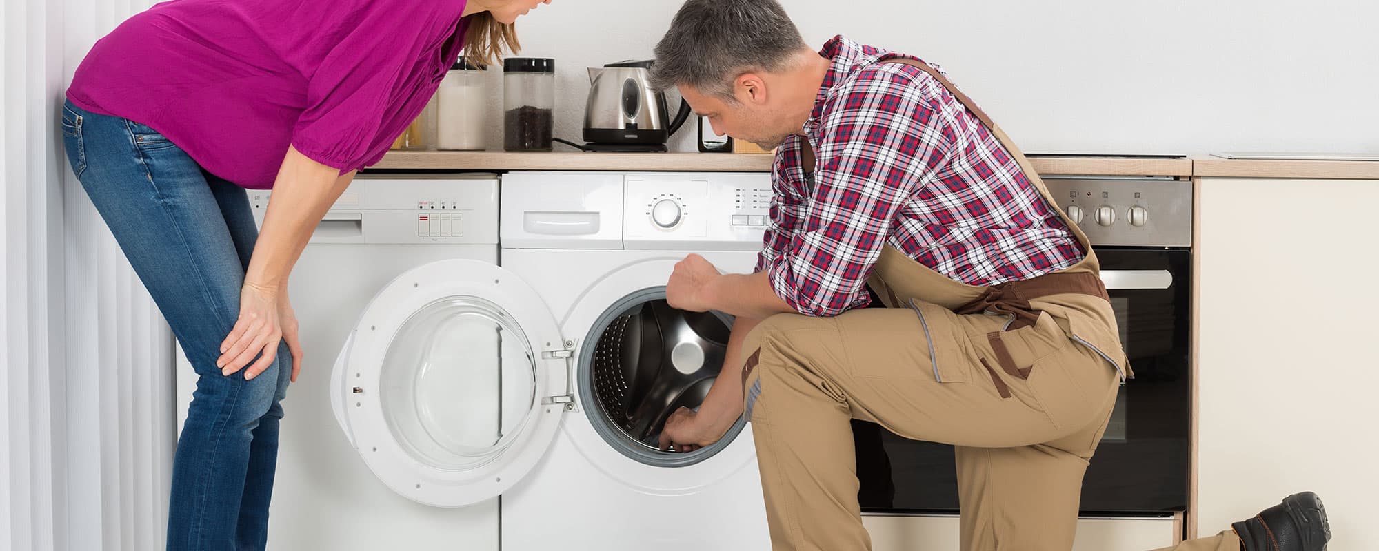 Woman Looking At Repairman Checking Washing Machine In Kitchen